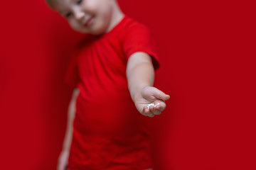 little sad boy kid hold a lot of pills in hands. red t-shirt, red background dangerous concept, tablets and baby