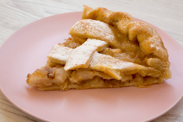 Slice of home-baked apple pie on a pink plate on a white wooden surface, side view. Close-up.