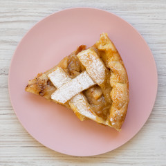 Slice of home-baked apple pie on a pink plate on a white wooden surface, top view. Overhead, from above, flat lay. Closeup.