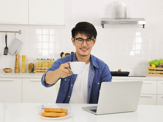 young handsome Asian man wearing eye glasses sitting at the kitchen table with computer laptop on it  smiling holding a cup of coffee and offer to the camera.