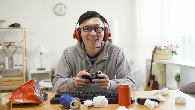 joyful man in headphones plays online video game in console looking camera with opened eye attention as computer screen monitor. happy excited asian male otaku stay at home on summer break eat snack