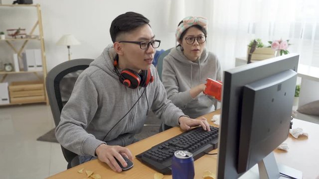 two asian couple intense play online video game on computer at home in dirty apartment eating junk food stay indoors on summer break. annoyed noisy woman talking point screen while boyfriend control