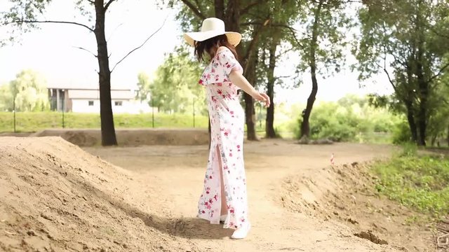 the girl walks through the sunny forest in a long white dress with red flowers. She is wearing a big straw hat and a girl is smiling at the camera.