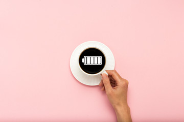 Female hand holding a white cup with black coffee on a pink background. Sign of a charged battery. Concept charge for the day. Flat lay, top view