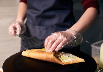 Step-by-step recipe for cooking dish. Chef cooking crepe on hot round cooktop. Skillful hands with black spatula rolling up big crepe greased with sauce. Traditional restaurant healthy food. Close up.