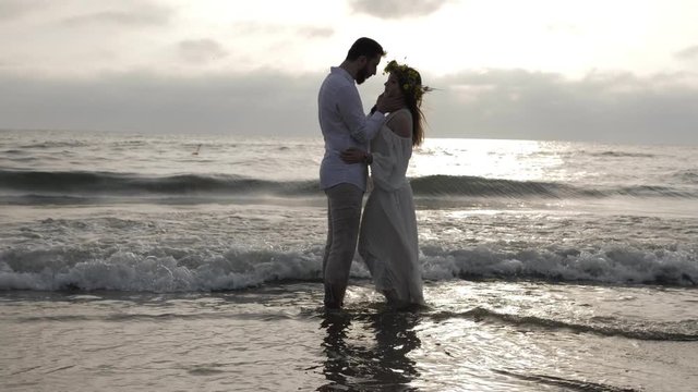 romantic couple silhouettes stand on sea water edge against foaming waves rolling on legs in summer evening slow motion