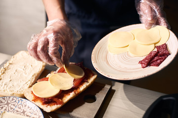 Cropped view of chef hands holding white plate with slices of cheese and sausage. Cook in gloves putting layers of pieces of ingredients on bread greased with red sauce. Traditional italian concept.