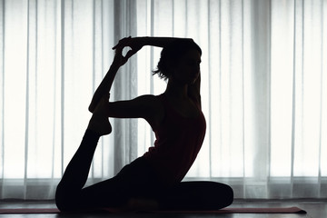Silhouette of sporty young woman practicing yoga indoors