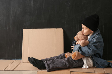 Homeless little girl with teddy bear sitting on floor near dark wall