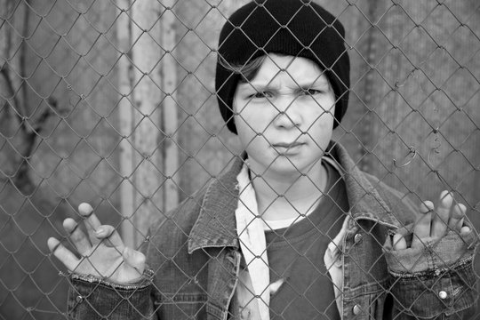 Homeless little boy standing behind mesh outdoors