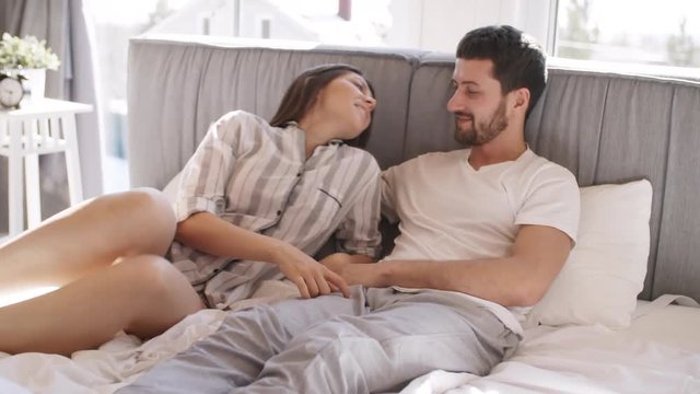 Handheld shot of young woman in pajama lying down on bed next to her boyfriend and kissing him on lips. Couple relaxing together in morning