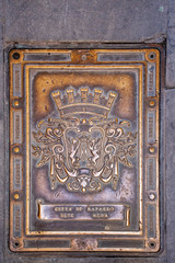 Close-up of a golden cast iron manhole cover in a street pavement with the symbols of the municipal coat of the city of Rapallo, rampant griffons holding the Marian acronym, Genoa, Liguria, Italy