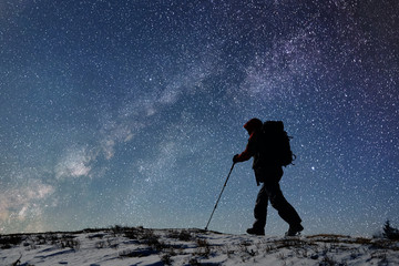 Silhouette of male tourist with backpack and trekking sticks going along snow-covered path high up...