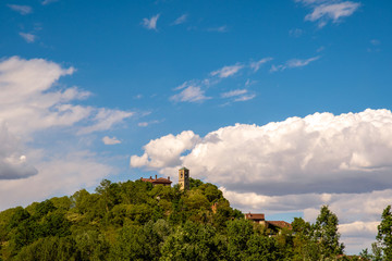 View of the top of a green hill with a bell tower among the trees and blue sky with white clouds, Piedmont, Italy