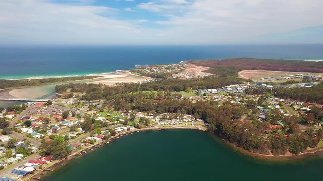 Aerial, reverse, drone shot, towards the village and nature on the coast of Burrill lake, on a sunny day, in New South Wales, Australia