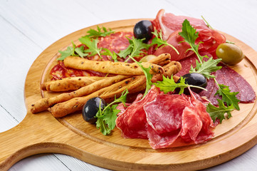 meat platter at white wooden table top with grissini, prosciutto, salami, sausage, and olives. close up