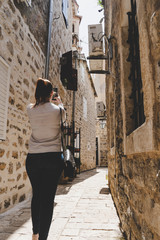 Young white girl taking pictures in the small narrow streets of the old town of Budva, Montenegro. Woman walking between medieval stone houses with smart phone