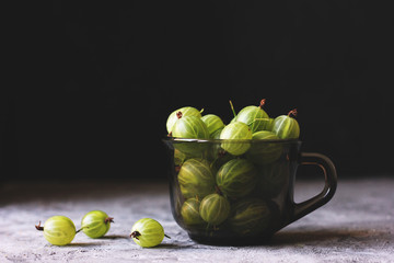 Dark and moody photo of juicy green gooseberries in a black glass cup standing on a grey background with a few berries lying around taken using natural light