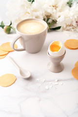 Soft-boiled egg with liquide orange yolk in ceramic egg cup, cup of coffee with ceramic spoon and thin crispy corn chips on background of beautiful white peony. Breakfast concept. Copy space.