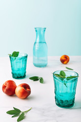 Two beautiful vintage turquoise glasses, a bottle with cold drink and ice cubes, and fruit decorated with fresh green mint leaves on white marble table. Copy space.