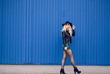 The girl in the black hat and leather jacket goes along the blue wall