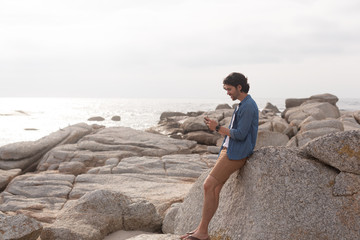 Caucasian man using mobile phone at beach while leaning on rock