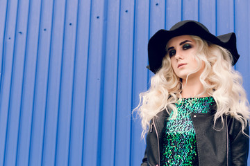 The girl in the black hat and leather jacket is standing and posing against the background of the blue wall