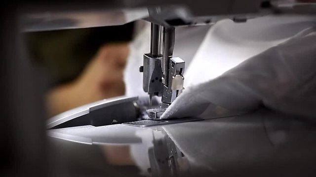 Close up view of presser foot making seam on white cloth with built-in hardware lamp turned on. Fingers straightening, drawing up fabric on darkened blurred background. Cutting off thread in the end
