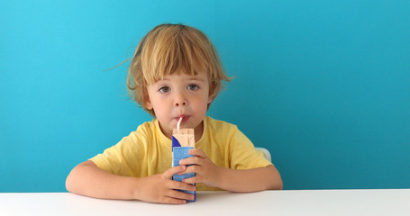Cute focused boy in yellow t-shirt drinking from carton box through straw sitting on blue background