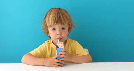 Cute focused boy in yellow t-shirt drinking from carton box through straw sitting on blue background