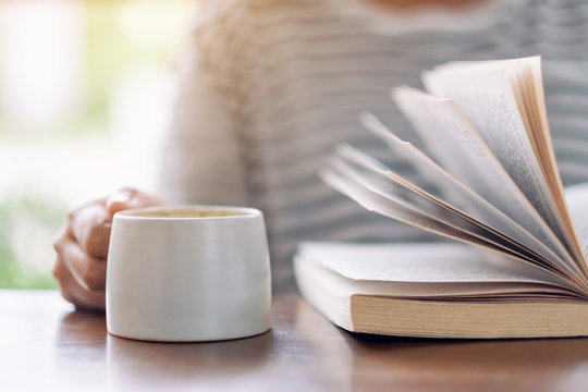 Closeup image of a woman holding and reading a book while drinking coffee on wooden table