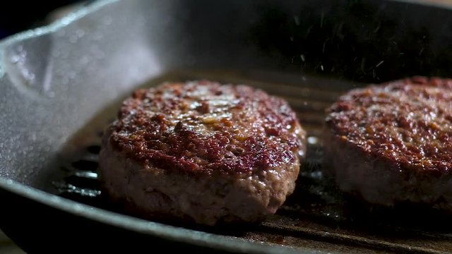 two meat fat juicy burgers with a hard crispy brown crust are fried on a hot frying pan with a non-stick grill. Boiling oil splashes heavily. Panoramic slow motion picture of the Cooking process in th