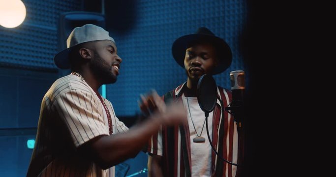 Two African American young guys rappers reading rap in the mic while recording a song together in the sound studio.