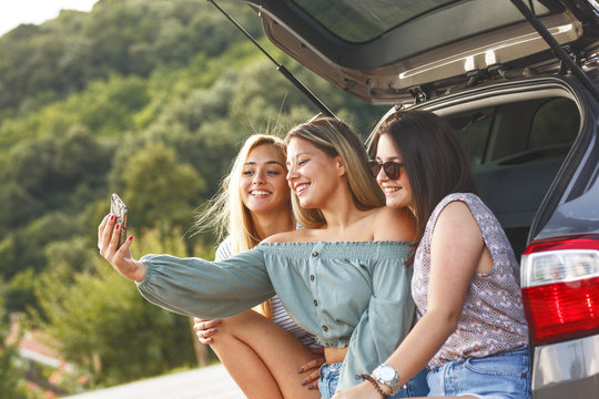 Group Of Female Best Friends On Travel . They Sitting On Car Trunk ,relaxing After Long Journey And Making Selfie.