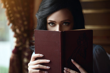 Close up portrait of young beautiful lady hiding the part of her face behind the open book