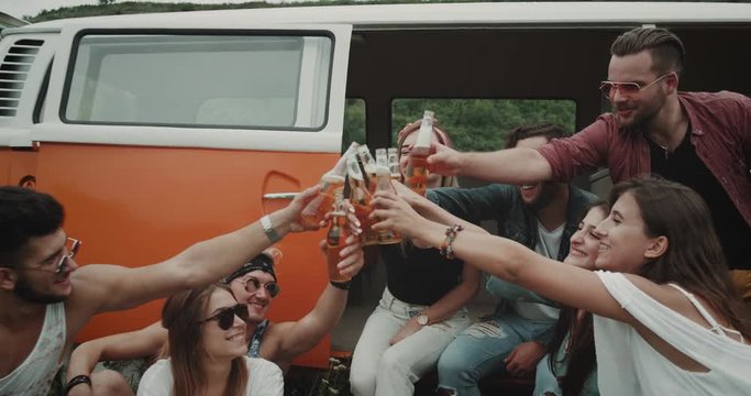 Boho stylish group of friends drinking beer and cheers at the picnic party , sitting behind the retro van