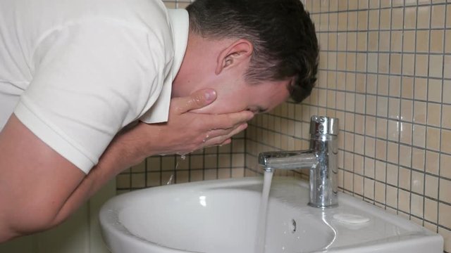 Morning hygiene in bathroom. Young man is washing his face early in morning in sink to wake up and get some energy, side view.