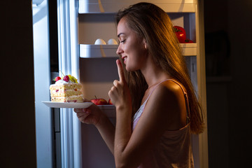 Hungry woman in pajamas eating sweet cake at night near fridge. Stop diet and gain extra pounds due...