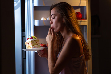 Hungry woman in pajamas enjoys sweet cake at night near refrigerator. Stop diet and gain extra pounds due to high carbs food and unhealthy eating