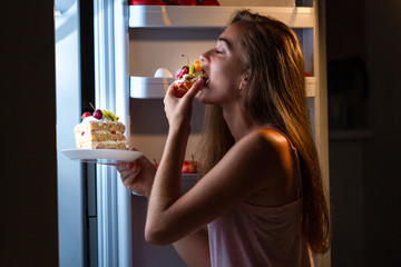 Hungry woman in pajamas eating sweet cakes at night near refrigerator. Stop diet and gain extra...