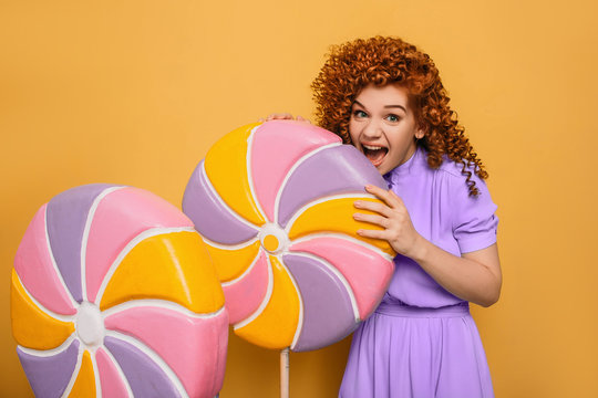 mmm yummy it is very sweet lollipop. Emotional curly haired woman bite off piece candy on yellow