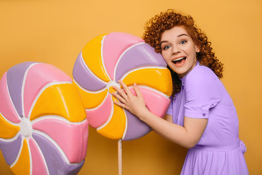 mmm yummy it is very sweet lollipop. curly red-haired woman hugging candy on yellow background
