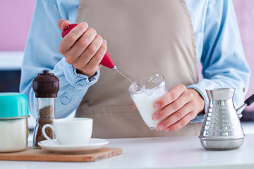 Woman in apron making coffee with using a milk frother at kitchen at home