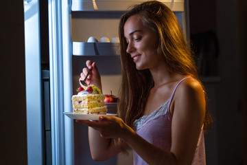 Hungry woman in pajamas eating sweet cake at night near fridge. Stop diet and gain extra pounds because of high carbs food and unhealthy night eating