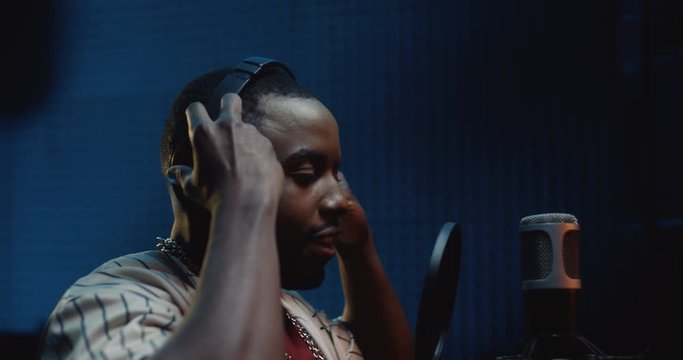 Close up of the young African American man in a hat taking on headphones and starting to record a song in the dark sound studio.