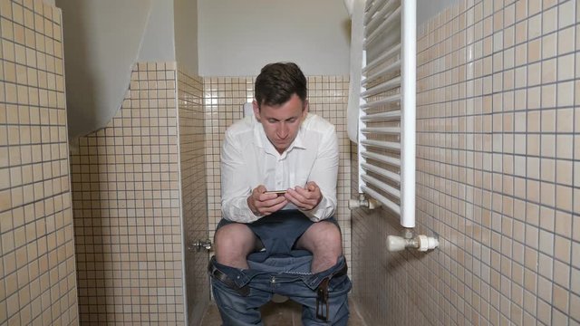Man holds his smartphone to pass the time sitting on toilet bowl in lavatory. He plays on the mobile phone.