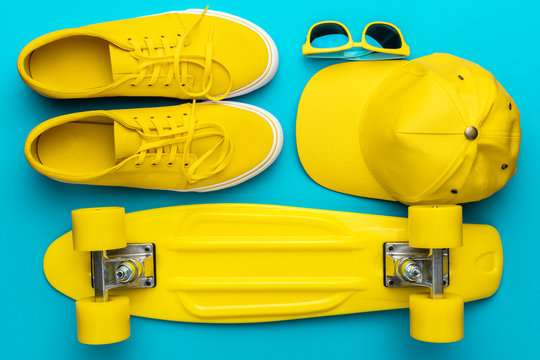 Top view of yellow modern teenage accessories in oder. Flat lay image of yellow baseball cap, sunglasses, sneakers, mini cruiser skateboard over blue turquoise background