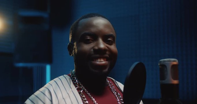 Portrait of the young African American handsome male singer smiling cheerfully to the camera while standing at the mic in the dark sound studio. Close up.
