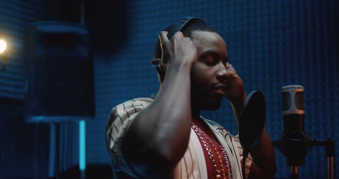 African American young stylish guy rapper taking on headphones and starting to record a song in the dark sound studio. Close up.