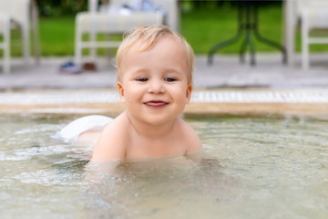 Happy little caucasian blond toddler boy swimming in wading pool on bright summer day at resort. Adorable baby enjoying outdoor water fun activities in shallow water. Children travel and holidays